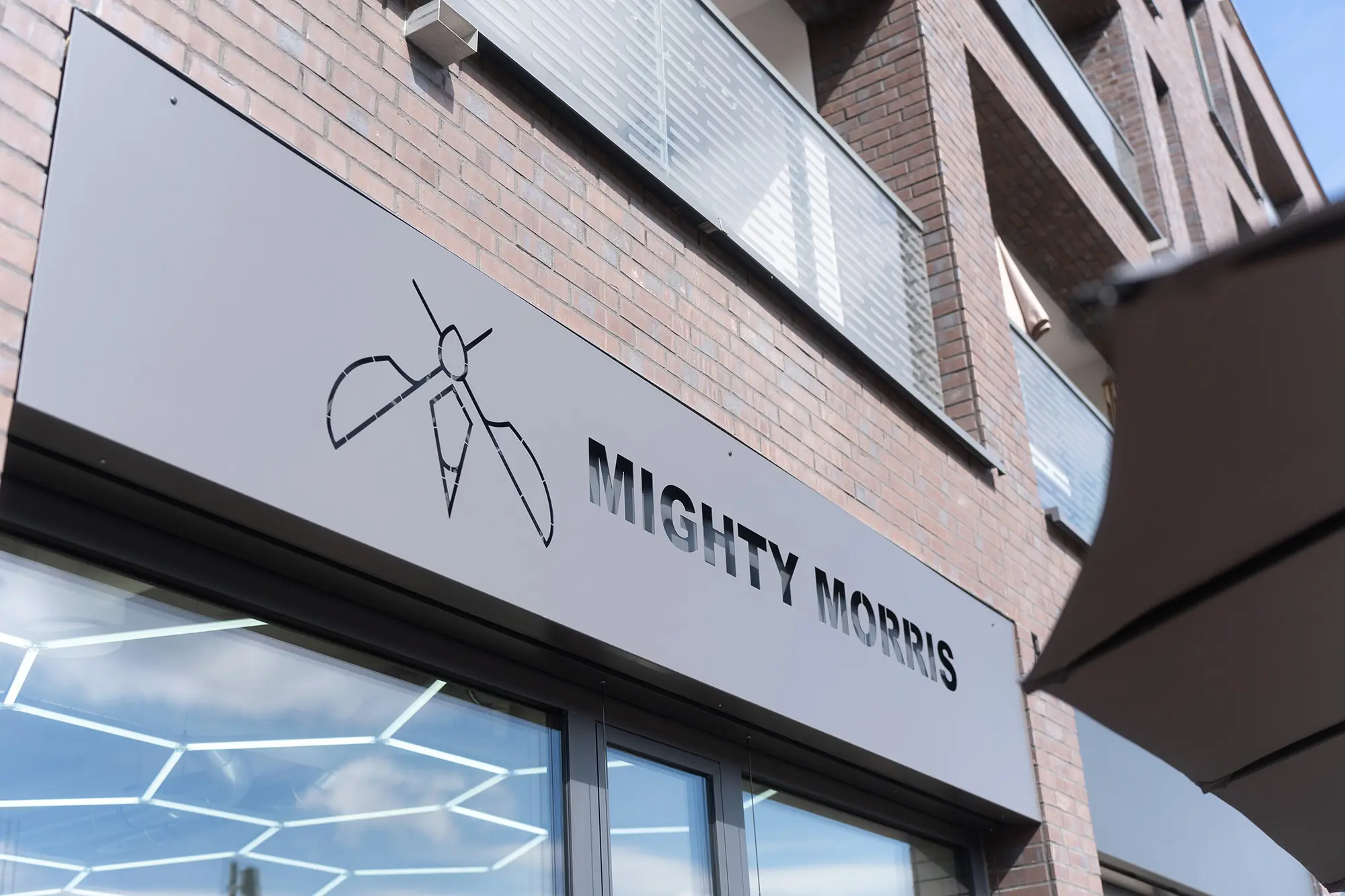 Das Ladenschild über dem Eingang: auf hellgrauem Grund das Mückenzeichen und der Schriftzug Mighty Morris, dahinter eine Klinkerfassade.