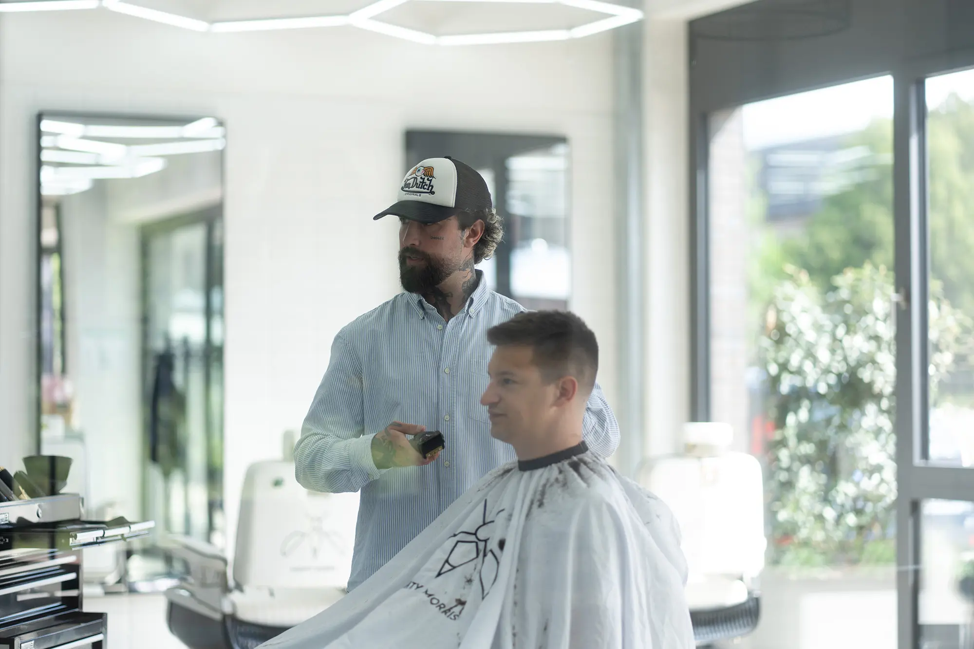 Ein Barbier mit Bart und Cap arbeitet mit der Maschine am Hinterkopf eines sitzenden Kunden. Der Umhang trägt das Mückenzeichen. Heller Raum, Tageslicht.